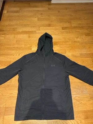 Svart vindjacka från Under Armour - Svart vindjacka från Under Armour med huva och hel dragkedja framtill. Jackan har två sidofickor och är tillverkad i ett lätt, syntetiskt material som passar perfekt för blåsiga dagar. Diskret logga på bröstet och sportig passform.