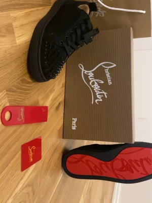 Svarta Loubs - Snygga svarta sneakers från Christian Louboutin med klassiska röda sulor och coola svarta nitar på tån. Skorna har snörning och är tillverkade i mocka och läder. Perfekta för dig som vill sticka ut med exklusiv design och edgy detaljer.