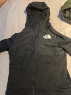 Svart vindjacka från The North Face - Svart vindjacka från The North Face med huva och hel dragkedja framtill. Jackan har två sidofickor och logga tryckt på både bröstet och ryggen. Perfekt för blåsiga dagar och enkel att matcha med olika outfits.