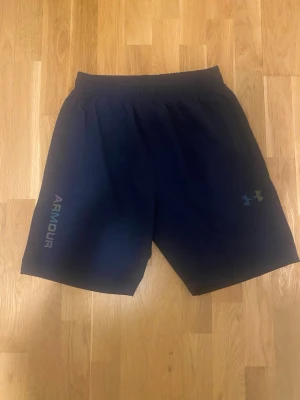 Blå träningsshorts från Under Armour - Mörkblå träningsshorts från Under Armour med elastisk midja och logga på ena benet. Shortsen har en enkel design med diskreta färgdetaljer baktill och är perfekta för sport eller gym. Lätta och bekväma att röra sig i.
