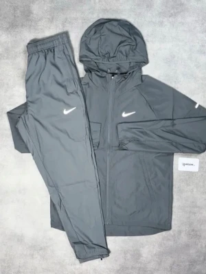 Grå Nike träningsset med huva - Snyggt grått träningsset från Nike med matchande jacka och byxor. Jackan har dragkedja, huva och vita Nike-loggor på bröst och byxa. Tillverkad i lätt och funktionellt material, perfekt för träning eller sportiga dagar.