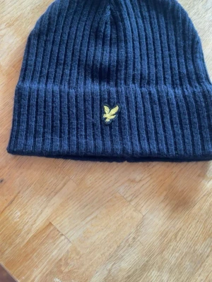 Svart ribbstickad mössa Lyle & Scott - Svart ribbstickad mössa från Lyle & Scott med klassisk gul logga framtill. Mjuk och stretchig modell som sitter skönt på huvudet. Perfekt accessoar för kyliga dagar.