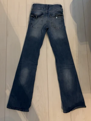 Blå bootcut jeans från Lindex  - Säljer ett par blå bootcut jeans från Lindex med klassisk femficksdesign och ljusa slitningar framtill och baktill. Jeansen har normal midja och stängs med knapp och dragkedja. Perfekta för dig som gillar en avslappnad och trendig look.