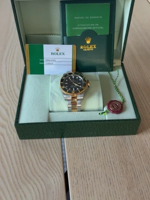 Lyxig svart/guld herrklocka - Säljer en lyxig  herrklocka med svart urtavla och gulddetaljer. Armbandet är i stål och guld, och klockan har en klassisk rund form med tydliga indexmarkeringar. Kommer med originalbox, certifikat och tillbehör.