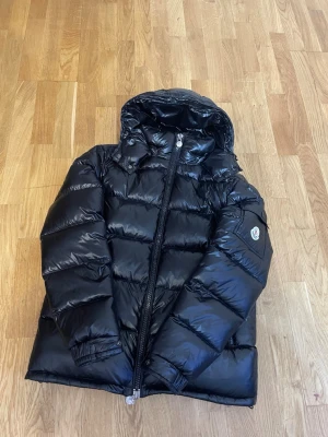 Svart dunjacka från Moncler - Snygg svart dunjacka från Moncler med glansig finish och klassisk pufferdesign. Jackan har huva, dragkedja framtill och Moncler-logga på ärmen. Insidan är grå och jackan är fylld med dun för extra värme under kalla dagar.  Storlek 3 passar både M och L