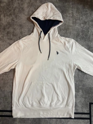 Vit hoodie från U.S. Polo Assn - Snygg vit hoodie från U.S. Polo Assn med mörkblå dragsnören och broderad logga på bröstet. Hoodien har känguruficka framtill och mörkblått innerfoder i huvan. Perfekt för en chill och stilren look.