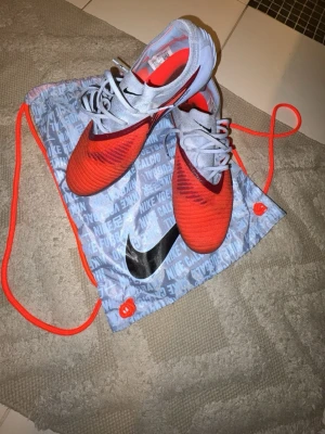 Nike röda och grå fotbollsskor - Säljer ett par snygga fotbollsskor från Nike i röd och ljusgrå färg. Skorna har en modern, strömlinjeformad design med snörning och tydlig Nike-logga. Perfekta för dig som vill sticka ut på planen och få bra grepp på konstgräs eller gräs.