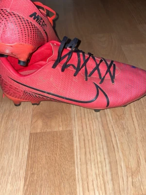 Nike Mercurial röda fotbollsskor - Säljer ett par röda Nike Mercurial fotbollsskor med svarta detaljer och snörning. Skorna har en låg profil, mönstrad ovansida och fasta dobbar för bra grepp på planen. Snygg design med svart Nike-logga på sidan. Perfekta för dig som vill ha snabbhet och kontroll på planen. Kan såklart tvätta skorna innan dom skickas 