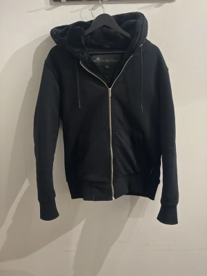 Moose Knuckles jacka - Snygg svart jacka från Moose Knuckles med dragkedja framtill, två stora fickor och mjuk fuskpäls i huvan. Klassisk passform och ribbade muddar vid ärmar och nederkant.