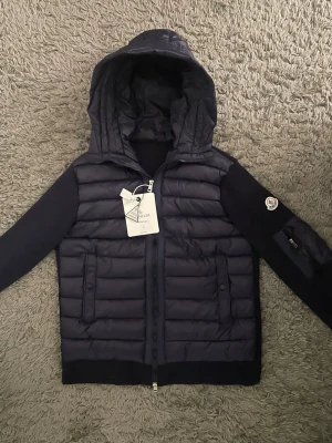 Mörkblå Moncler cardigan med luva  - Snygg mörkblå  Moncler cardigan med quiltad front och huva. Jackan har stickade ärmar och muddar, två fickor med knappstängning och Moncler-logga på ena ärmen. Perfekt för kalla dagar och riktigt trendig look.