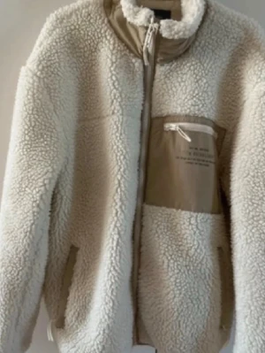 Crémevit teddyjacka från Bershka - Säljer en teddyjacka från Bershka med dragkedja framtill och hög krage. Jackan har en kontrasterande beige bröstficka med dragkedja och två sidofickor. Materialet är mjukt och fluffigt, perfekt för kyliga dagar. Den har ett hål i fickan men det går lätt sy ihop! Kan göra det innan jag postar för 30kr extra