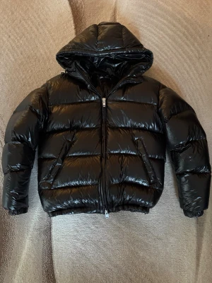 Svart pufferjacka från Zara  - Snygg svart pufferjacka från Zara med glansigt material och stor huva. Jackan har dragkedja framtill och två snedställda fickor. Perfekt för kalla vinterdagar och ger en cool look. Passar dig som vill ha en varm och trendig jacka.