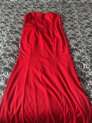 Röd axelbandslös långklänning - Snygg röd axelbandslös långklänning med figurnära passform. Klänningen har en hjärtformad byst och är perfekt för dig som vill sticka ut. Materialet känns mjukt och följsamt, vilket gör den bekväm att bära hela kvällen. Den har en fått ett svart sträck samt som en litet hål men det är längst ner på klänningen vilket gör att det inte syns när man har på sig den