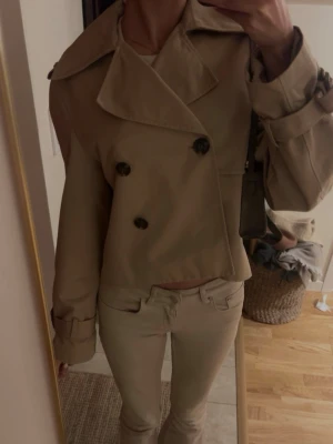 Kort beige trenchcoat med stora slag - Stilren kort trenchcoat i beige med dubbelknäppning och stora svarta knappar. Jackan har breda slag, klassiska axelklaffar och justerbara ärmslut. Perfekt för en trendig och tidlös look. Materialet är troligtvis bomull eller bomullsblandning.