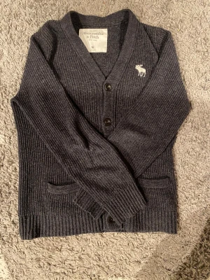 Mörkgrå kofta Abercrombie & Fitch M - Snygg mörkgrå ribbstickad kofta från Abercrombie & Fitch i storlek M. Koftan har v-ringning, två framfickor, knappar och en broderad vit älg på bröstet. Perfekt för lager-på-lager och chill stil.
