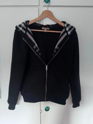 Svart hoodie från Burberry med dragkedja - Svart hoodie från Burberry med klassiskt rutigt foder i huvan och dragkedja framtill. Broderad Burberry-logga på bröstet. Mjuk bomullskänsla och två fickor framtill. Perfekt för en stilren och avslappnad look.