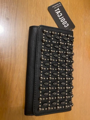 Svart plånbok med nitar från Coolcat - Snygg svart plånbok från Coolcat med coola silverfärgade nitar och små döskallar på framsidan. Plånboken är gjord i konstläder och har flera fack för kort, sedlar och mynt med dragkedja. Perfekt accessoar för dig som gillar edgy detaljer.
