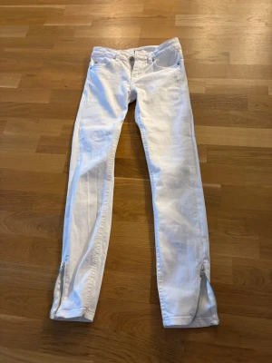 Vita jeans med dragkedjor nertill - Säljer ett par vita jeans med smal passform och dragkedjor vid bensluten. Klassisk femficksmodell och knappgylf. Perfekta för dig som vill ha en clean och stilren look.