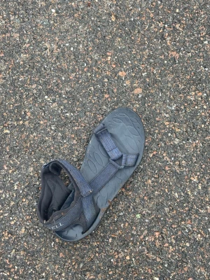 Svart sportig sandal från Merrell - En svart sandal från Merrell med justerbara kardborreband och robust sula med mönster. Perfekt för dig som gillar en sportig och funktionell stil. Tillverkad i slitstarkt syntetmaterial och textil för extra komfort. ORGINAL PTIS 200