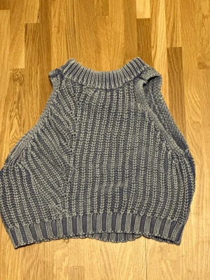Gråblå stickad väst med fläta - Säljer en gråblå stickad väst i grov ribbstickning med breda ärmhål och hög ribbad hals. Framsidan har en snygg fläta som detalj. Perfekt att styla över en skjorta eller t-shirt för en trendig lager-på-lager-look.