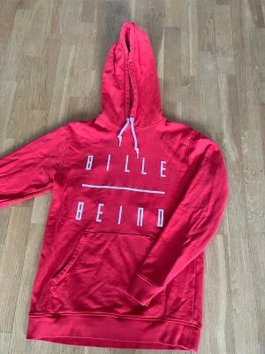 Röd hoodie från Billebeino - Snygg röd hoodie från Billebeino med vit text på bröstet och en stor magficka. Hoodien har dragsko i huvan och ribbade muddar vid ärmslut och nederkant. Perfekt för en avslappnad streetwear-look.