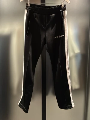 Palm Angels svarta mjukisbyxor - PRISET KAN DISKUTERAS!!!   Svarta mjukisbyxor från Palm Angels med vita sidoränder och logga tryckt på vänster ben. Byxorna har elastisk midja och raka ben. Perfekta för en avslappnad streetwear-look. Hör av er vid frågor eller prisförslag! 👍