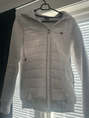 Vit quiltad hoodiejacka från Tommy Hilfiger - Snygg vit hoodiejacka från Tommy Hilfiger med quiltade paneler framtill och dragkedja hela vägen. Jackan har huva, ribbade muddar och två sidofickor. Perfekt för dig som gillar en clean och sportig look.