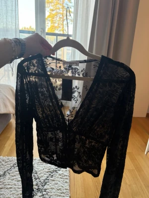 Svart spetsblus från venderbys  - Snygg svart blus från venderbys i transparent spets med blommigt mönster. Modellen är croppad med lång ärm och djup v-ringning. 