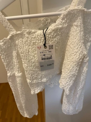 Vit offshoulder blus med struktur - Trendig vit offshoulder blus med långärmad design och strukturerat tyg. Blusen har volangdetaljer upptill och är tillverkad i ett lätt, luftigt material som ger en romantisk känsla. Perfekt för dig som vill ha en söt och modern look.