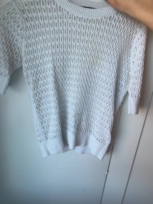 Vit stickad kortärmad tröja - Säljer en vit stickad tröja med korta ärmar och snyggt hålmönster. Tröjan har ribbstickad halsringning, ärmslut och nederkant. Perfekt för dig som gillar en clean och stilren look.