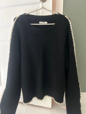 Svart stickad tröja med vita detaljer - Svart stickad tröja från Indiska med vita virkade detaljer längs ärmar och nederkant. Tröjan har rund halsringning, långa ärmar och vågig kant nertill för en cool touch. Perfekt för dig som gillar en enkel men unik stil. Är i storlek M men skulle säga att den är lite overzised 