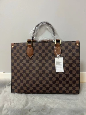 Brun rutig handväska med skinnhandtag - Snygg handväska i brunt med rutigt mönster och detaljer i mörkbrunt skinn. Väskan har rejäla handtag med guldfärgade metalldetaljer och en boxig, rektangulär form. Perfekt för dig som gillar klassisk och stilren design.