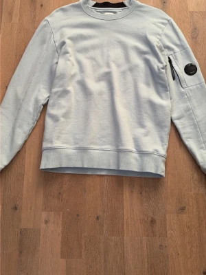 Ljusblå sweatshirt från C.P. Company - Ljusblå sweatshirt från C.P. Company med rund halsringning. Tröjan har en ikonisk linsdetalj och dragkedjeficka på ena ärmen. Perfekt för en avslappnad och stilren look.