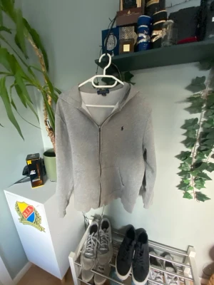 hoodie  Polo Ralph Lauren - XL i barnstolen! Sitter som XS/S ! Snygg grå hoodie från Polo Ralph Lauren med dragkedja framtill och klassisk huva. Tröjan har en liten broderad logga på bröstet och är gjord i mjukt bomullsmaterial. Perfekt för en avslappnad och stilren look.