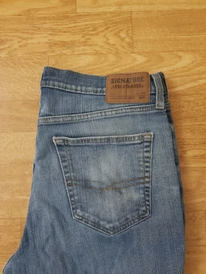  Levi's Signature blåa jeans  - Ett par klassiska blå jeans från Levi's Signature med femficksdesign och snygga slitningar. Jeansen har en bakficka med diskret broderi och läderpatch med logga i midjan. Perfekta för en avslappnad och trendig look.