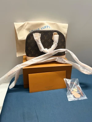  Alma BB Monogram - Snygg handväska  i modellen Alma BB. Väskan är i klassiskt brunt monogram canvas med beige läderdetaljer och guldfärgade metalldelar. Kommer med dustbag, ask och axelrem. Perfekt för dig som gillar lyxiga accessoarer och tidlös design.