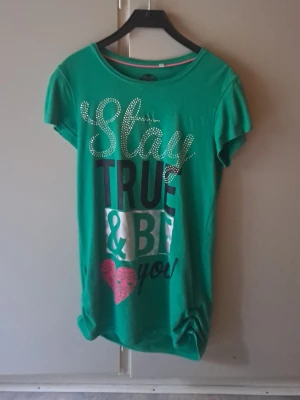 Grön t-shirt med tryck från C&A - Grön t-shirt från C&A here+there med texten 'Stay True & Be You' i vitt, svart och silver samt ett rosa hjärta. T-shirten har korta ärmar och rund hals. Dekorerad med glittriga detaljer på texten. 2000s & early 2010s vibe. Vid sidan har den som nån detalj som gör att den är tajtare o man får därmed mer form där nere. Storlek 164 jag beskriver som XS/S. Inga defekter