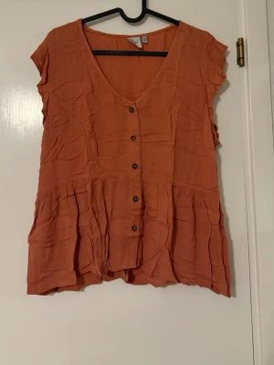 Rostfärgad blus från ICHI - Superfin rostfärgad blus från ICHI med knappar framtill och v-ringning. Blusen har korta ärmar och ett avslappnat, lite rynkat fall. Perfekt att styla med jeans eller kjol. Materialet känns lätt och luftigt.