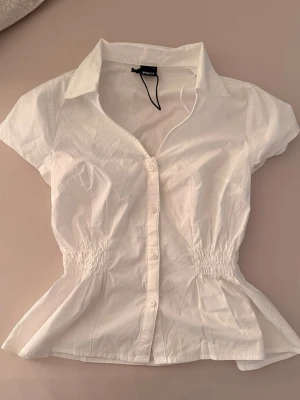 Vit kortärmad blus med krage - Snygg vit blus från New Yorker med korta ärmar, klassisk krage och knäppning framtill. Blusen har smock i midjan för en figurnära passform och är tillverkad i ett lätt bomullstyg. Perfekt för en fräsch och stilren look. Jätte söt blus nu till sommaren😍 helt ny och oanvänd bara prislappen av.