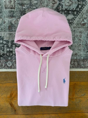 Ljusrosa Polo Ralph Lauren Fleece Hoodie - Storlek L - Hej! 👋 Säljer en riktigt snygg ljusrosa Polo Ralph Lauren hoodie i storlek L med mörkblå pony-logga. 🌸🐎 Tröjan är i mycket gott skick och känns superfräsch. Det finns 4 st minimala prickar på ena ärmen som knappt syns vid användning (se sista bilderna). 100% äkta med originaltvättlappar kvar och en fullt fungerande QR-kod för superenkel verifiering ✅ Perfekt färgklick till garderoben! Fraktar snabbt! 💬 🚛📦✨ 