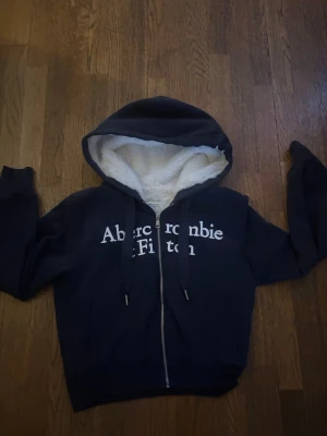 Mörkblå hoodie Abercrombie & Fitch - Mysig mörkblå hoodie från Abercrombie & Fitch med vit teddyfoder i huvan och dragkedja framtill. 