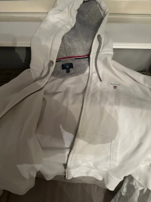Vit hoodie från Gant med dragkedja - Vit hoodie från Gant med dragkedja framtill och justerbar huva med grå insida. Klassisk logga broderad på bröstet och fickor på sidorna. Perfekt för en avslappnad och stilren look.