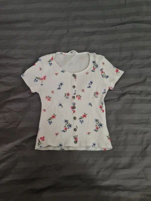 Vit blommig ribbad topp med knappar - Supersöt vit ribbad topp med korta ärmar och blommigt mönster i rosa och blått. Toppen har rund halsringning och knappar framtill, perfekt för en fräsch och somrig look.