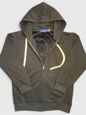Svart hoodie från Polo Ralph Lauren - Svart hoodie med dragkedja från Polo Ralph Lauren. Har vit dragsko i huvan och röd broderad logga på bröstet. Mjuk insida och ribbade muddar vid ärmslut och nederkant. Perfekt för en chill och stilren look.