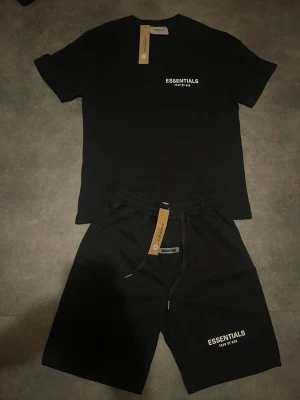 Svart t-shirt från Essentials - Svart t-shirt från Essentials med vit logga tryckt på bröstet. Klassisk rund halsringning och korta ärmar. Tillverkad i mjuk bomull för en skön och avslappnad känsla. Perfekt för en clean och stilren look.
