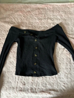 Svart offshoulder topp med knappar - Trendig svart ribbad topp med offshoulder-design och långa ärmar. Toppen har snygga knappar framtill och en figurnära passform som ger en cool och stilren look. Perfekt att matcha med jeans eller kjol för en modern outfit.