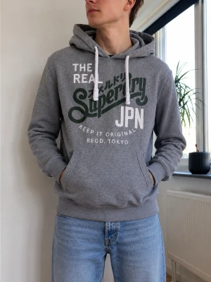 Grå hoodie från Superdry - Snygg grå hoodie från Superdry med stor logga och text i vitt och mörkgrönt på bröstet. Klassisk känguruficka framtill, vita dragsnören i huvan och mjukt material. Perfekt för en avslappnad streetwear-look.
