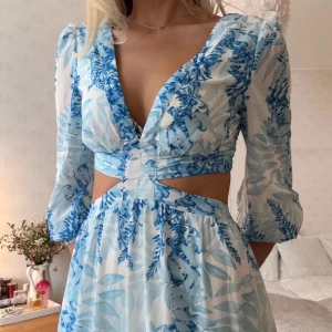 Långklänning - Säljer en superfin långklänning i blått och vitt med blommigt mönster. Klänningen har v-ringad hals, långärmade puffärmar och snygga cut-outs vid midjan.