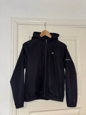 Windbreaker från Lacoste - Blå windbreaker från Lacoste med huva och dragkedja framtill. Jackan har diskret Lacoste-logga på bröstet och märkesnamnet på ärmen. Tillverkad i lätt och tåligt material, perfekt för blåsiga dagar.