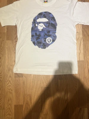 Vit BAPE t-shirt med blå camo-tryck - Vit t-shirt från BAPE med det ikoniska apansiktet i blått camo-tryck på bröstet. Klassisk passform med korta ärmar och rund hals. Tillverkad i mjuk bomull och har en liten BAPE-tag på ärmen. Perfekt för dig som gillar streetwear.